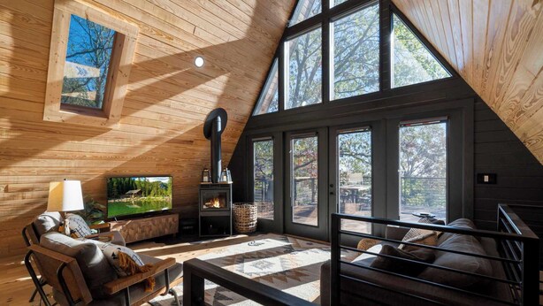 Un acogedor interior de cabaña con chimenea, paredes de madera, un sofá cómodo y ventanas amplias que ofrecen vistas al exterior.