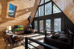 Un acogedor interior de cabaña con chimenea, paredes de madera, un sofá cómodo y ventanas amplias que ofrecen vistas al exterior.