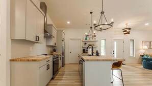 Une cuisine moderne avec des meubles blancs, un plan de travail en bois et un îlot central. Des luminaires suspendus sont placés au-dessus de l’îlot, et un four encastré est intégré.