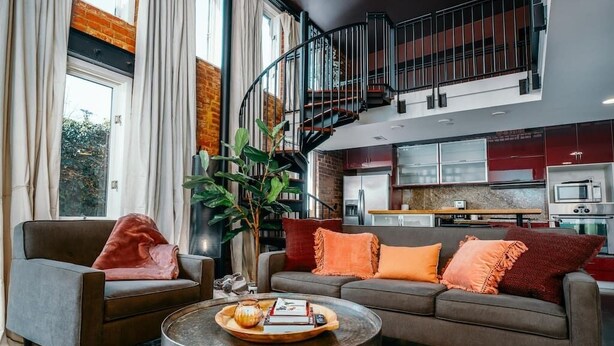 A modern living room with a sofa, armchairs, and a coffee table. A staircase leads to an upper level. - The Loft on Campus Su (Fayetteville)