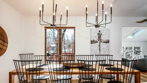 Un coin repas avec une table en bois et des chaises noires, un lustre et une grande fenêtre donnant sur les arbres.