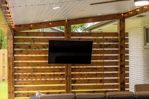 An outdoor seating area with a flat-screen TV mounted on a wooden slatted wall, surrounded by cushioned seating. - Porch on 3rd, Bentonville, bikable to DT and Coler (Bentonville)
