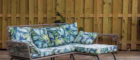 A wicker sectional sofa with a tropical leaf pattern, positioned against a wooden fence.