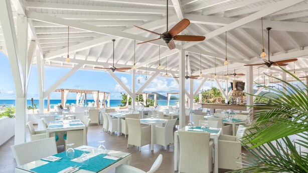 Un amplio comedor al aire libre con mobiliario de mimbre blanco y ventiladores de techo, con vista al océano.