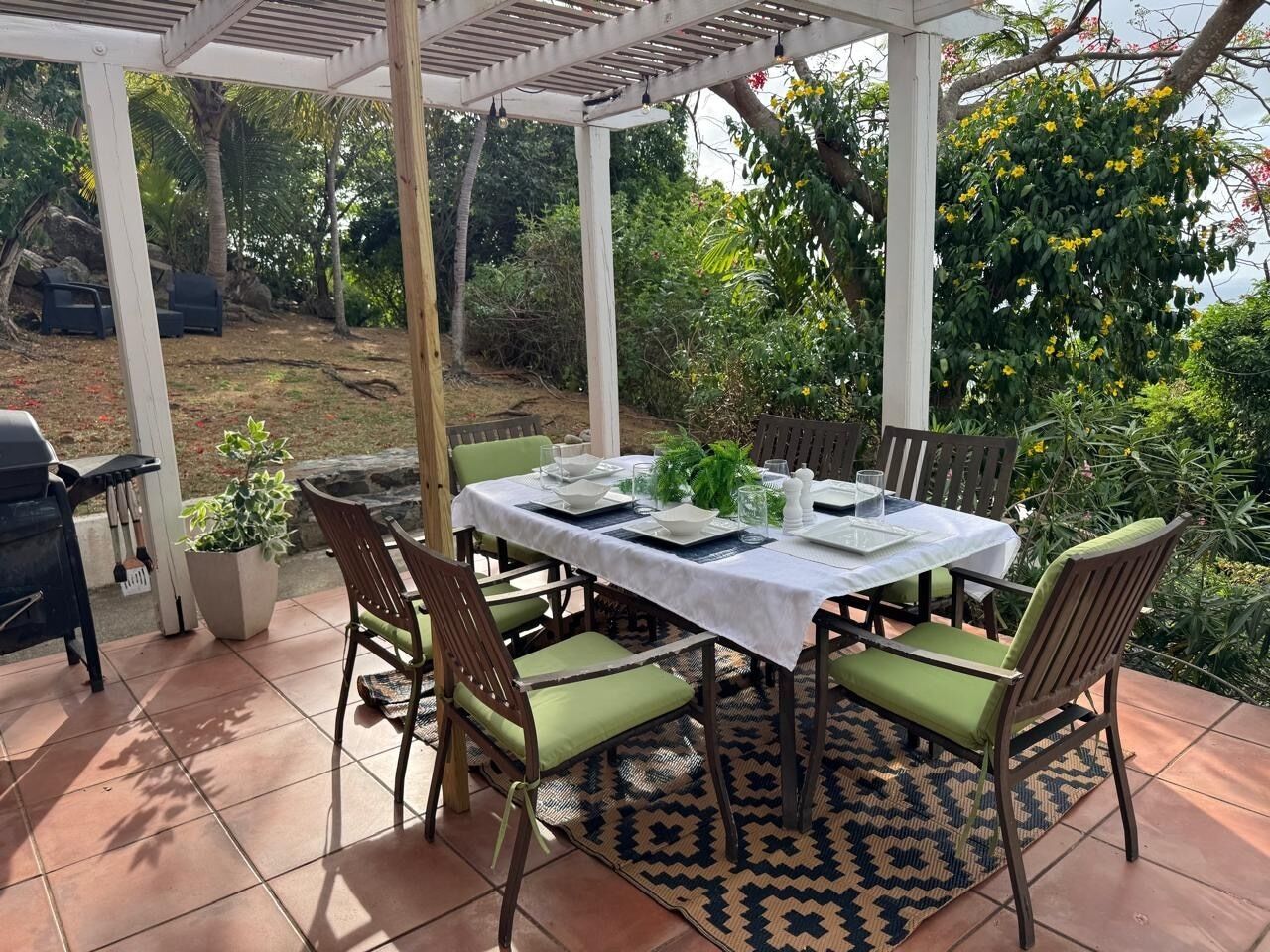 A covered patio with a table set for four, surrounded by chairs with green cushions, a patterned rug, and a grill in the background.