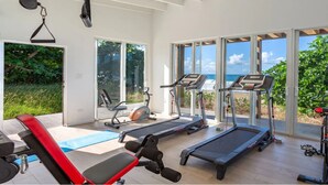 Un gimnasio con cintas de correr, bicicletas estáticas y máquinas de pesas, con amplias ventanas y vistas al océano.