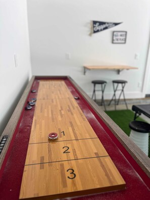 A shuffleboard table with numbered scoring areas, a small table, and two stools in the background. - Sapphire Lodge at Bella Vista (Bella Vista)