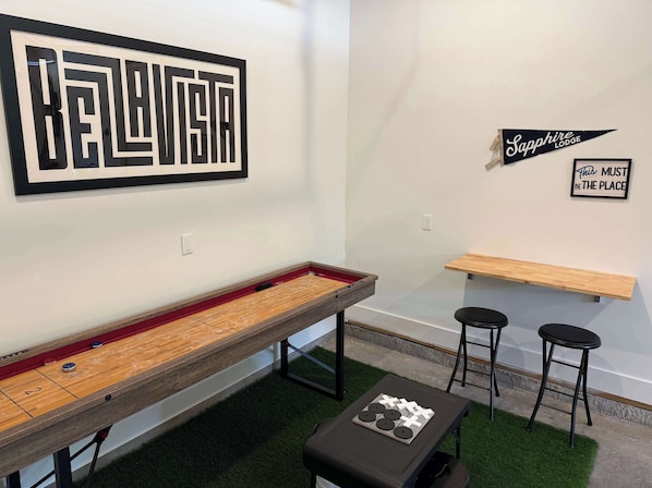 A room with a shuffleboard table, two bar stools, a small table with a board game, and a wall with a 'Sapphire Lodge' sign and a motivational quote.