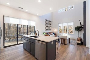 A modern kitchen with a large island, sliding glass doors to a deck, and a dining area with a view of trees. - Sapphire Lodge at Bella Vista (Bella Vista)