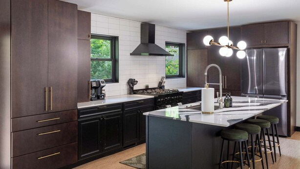 A modern kitchen with dark cabinetry, a central island with stools, a stainless steel refrigerator, and a pendant light fixture.