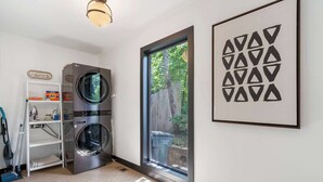 A laundry room with two stacked washers and dryers, a window with a view of a wooden fence and greenery, and a framed geometric art piece on the wall.