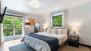 A bedroom with a bed, bedside lamps, a window with curtains, a floor lamp, and a view of greenery outside.