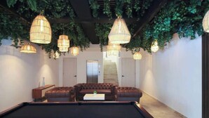 A pool table with a brown leather sofa and a coffee table in a room with hanging lights and green foliage. - Rooftop Pool + Gym + Laundry + Coworking + Cinema (Tulum)