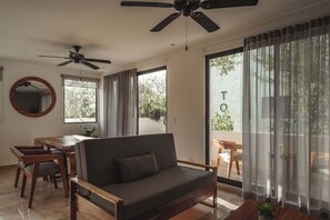 A modern living room with a sofa, dining table, chairs, ceiling fans, and large windows with curtains.