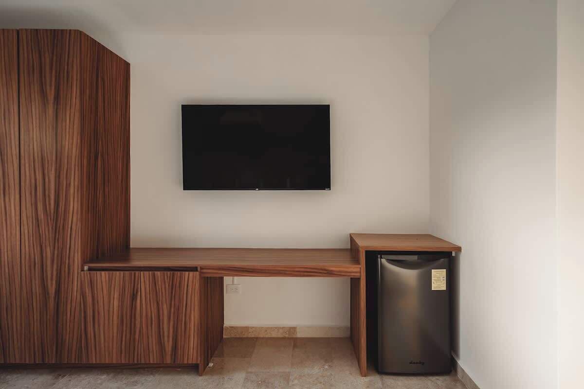A room with a wooden desk, a black television mounted on the wall, and a black trash bin.
