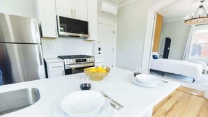 Une cuisine moderne dotée d’appareils électroménagers en acier inoxydable, un îlot central avec un saladier rempli de fruits, et un coin repas avec une table blanche mise pour deux personnes.