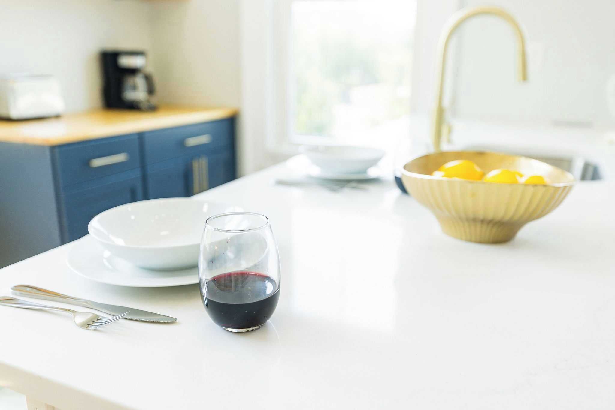 Uma bancada de cozinha com um copo de vinho tinto, uma tigela de limões e uma cafeteira ao fundo.