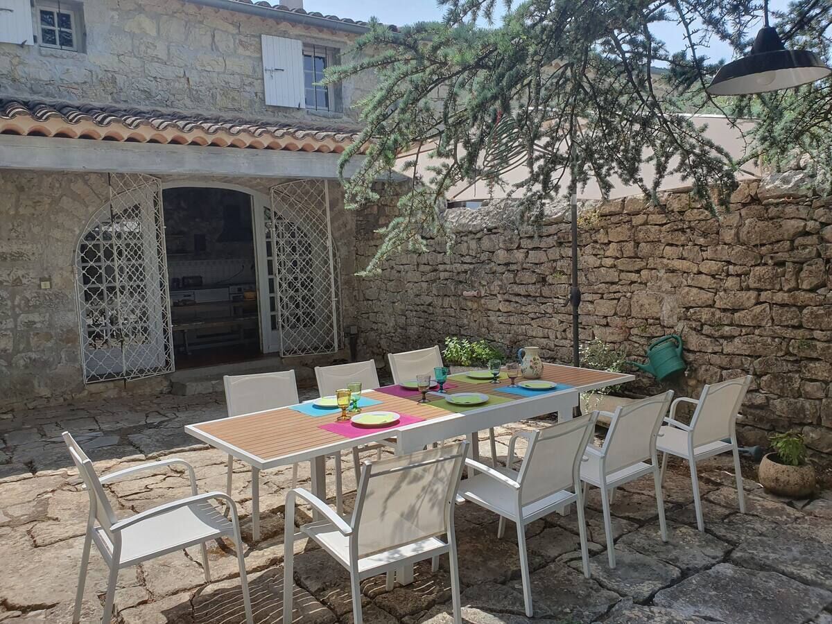 An outdoor patio with a stone wall, a wooden table, white chairs, and a metal gate.