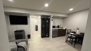 Uma sala de estar moderna com um sofá cinza, uma televisão de tela plana e uma área de cozinha com mesa e cadeiras.