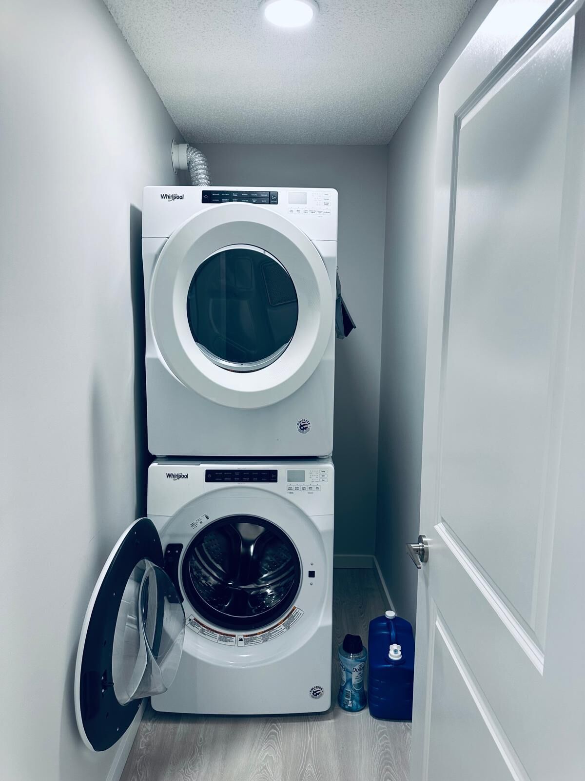 A laundry room with two stacked front-loading washing machines and a blue detergent bottle on the floor.