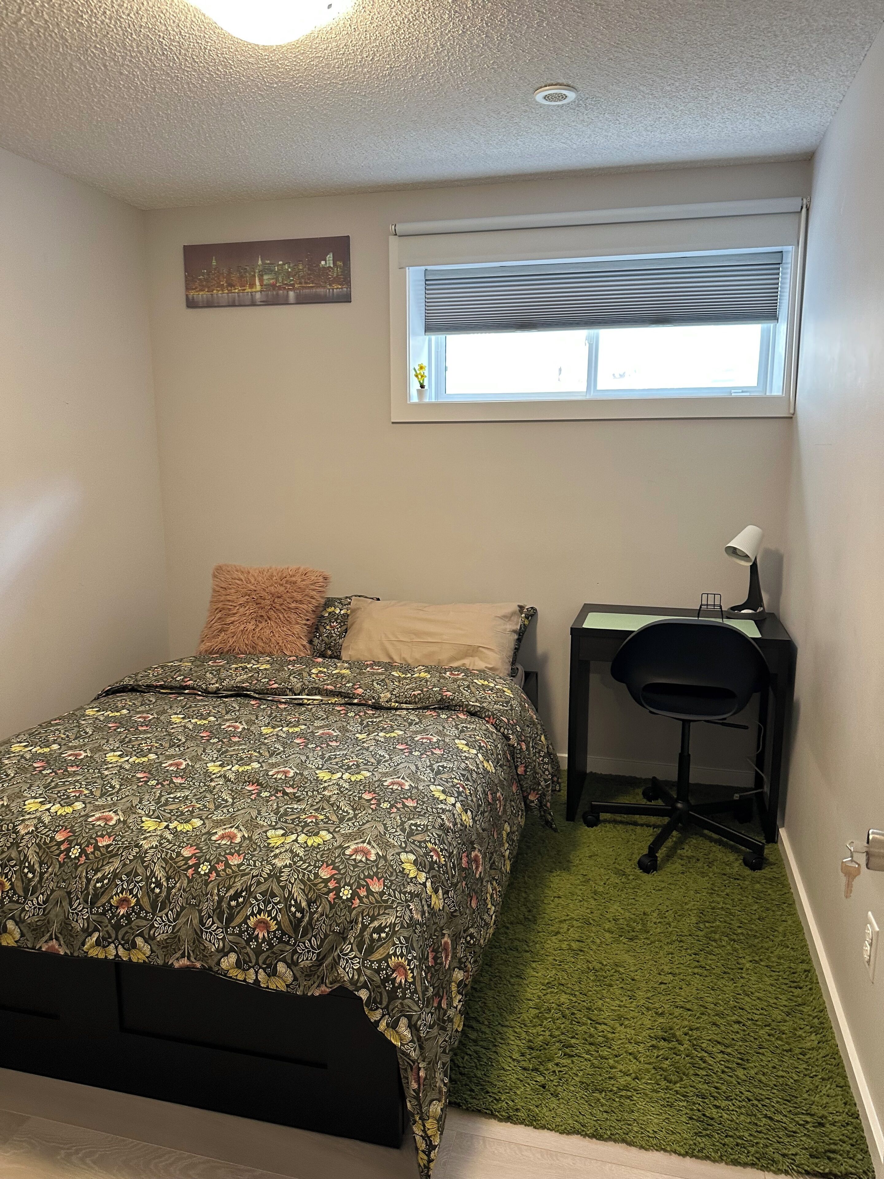 A bedroom with a bed, a desk, a chair, a window with blinds, and a small picture on the wall.