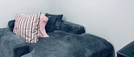 Ein modernes, minimalistisches Schlafzimmer mit einem dunkelgrauen Schlafsofa, einem Streifen-Kissen und einer rosafarbenen Tagesdecke. Zwei gerahmte Bilder hängen an der Wand.