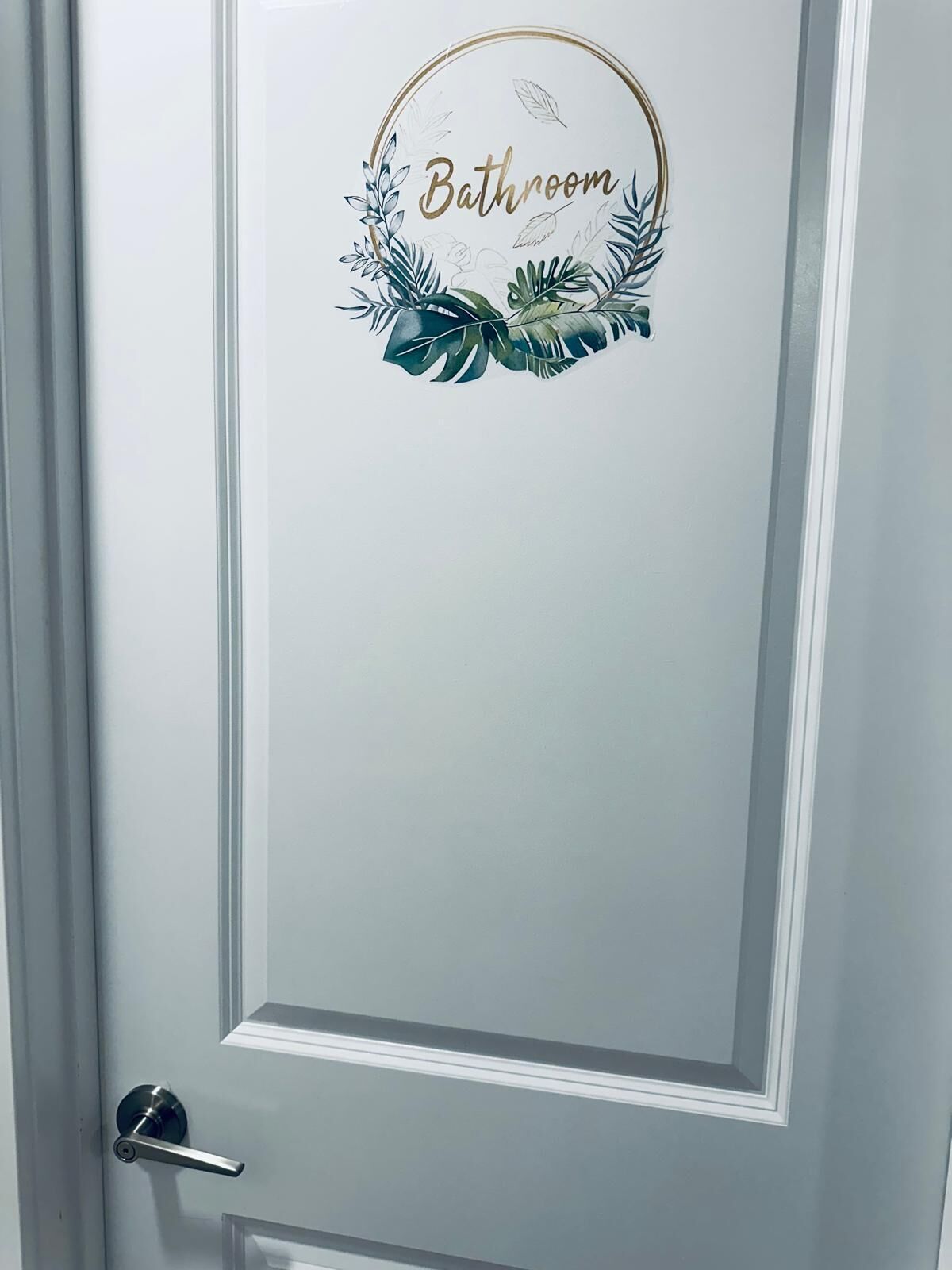 Una puerta con un adhesivo circular que dice "Baño", rodeada por una guirnalda decorativa.