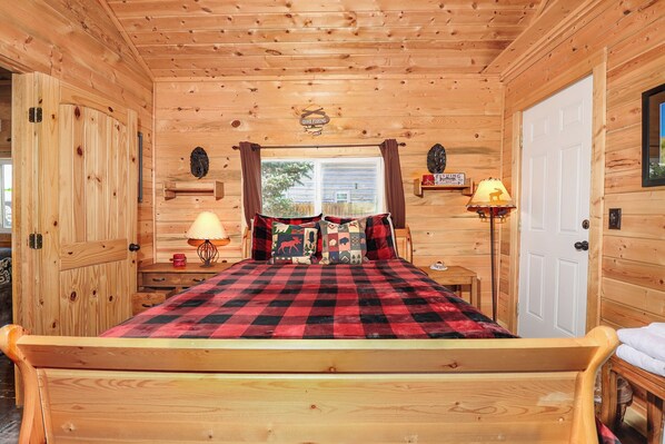 A wooden cabin bedroom with a bed, two bedside tables, a lamp, and a window with curtains.