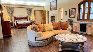 Een ruime woonkamer met een grote bank, een ronde salontafel en een houten vloer. Achtergrond is een bed met gordijnen en een open haard.