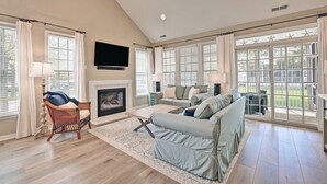 A living room with a fireplace, a sofa, a TV, and large windows. - Incredible 4BD Bear Trap Dunes Vacation Retreat! End Unit Townhome with Community Pool, Free WIFI, Golf, Beach Shuttle, Tennis, Pickleball and more! (Ocean View)