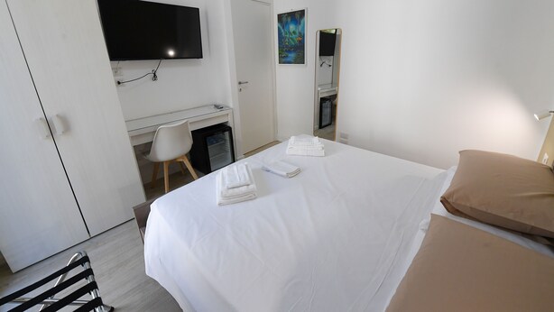 A hotel room with a bed, a desk with a chair, a television, and a painting on the wall. - Scilla SeaFront Rooms (Scilla)