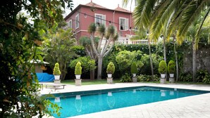 A swimming pool with a tiled deck, surrounded by potted plants and a house in the background. - Suites de Franchy (La Orotava)