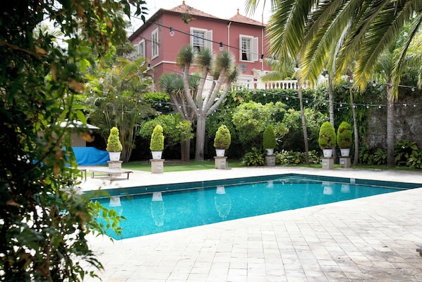 Une piscine avec une terrasse carrelée, entourée de plantes en pot et d’une maison en arrière-plan.