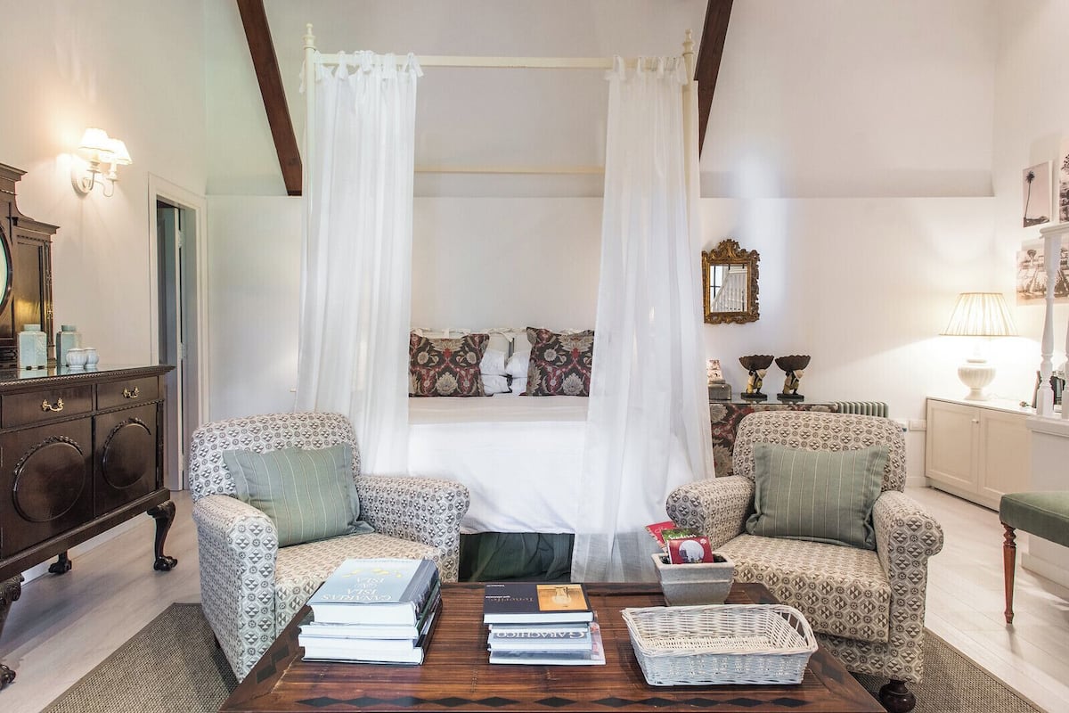 A bedroom with a canopy bed, two armchairs, a wooden coffee table with books, and a small side table.