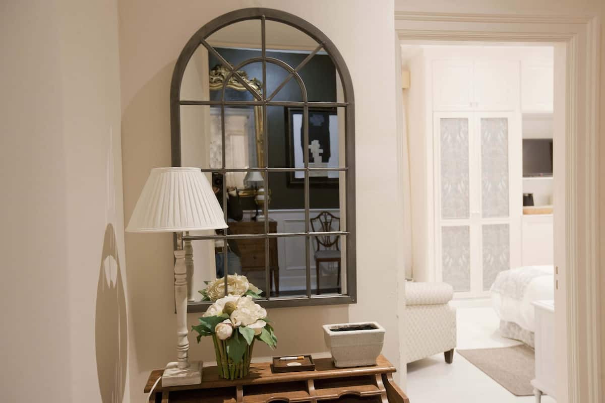 A bedroom with a large arched window, a bedside table with a lamp, a vase of flowers, and a bed with a white headboard.