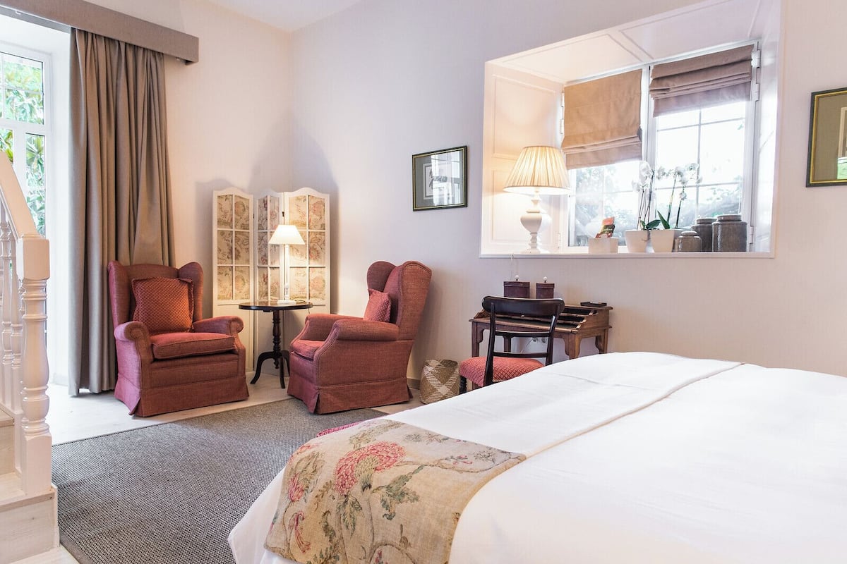 A hotel room with a large bed, two armchairs, a small table with a lamp, a piano, and a window with curtains.