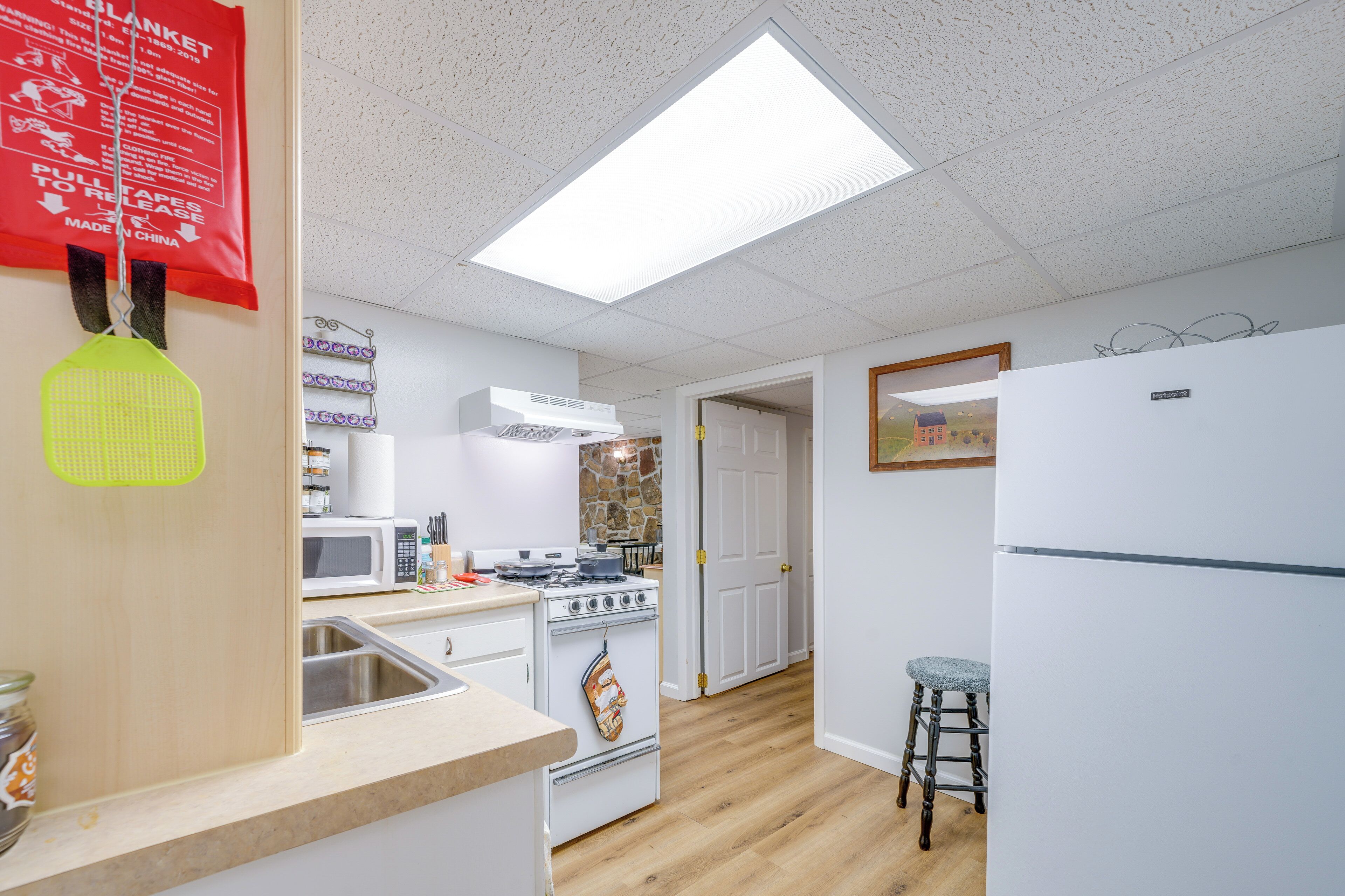 A kitchen with white appliances, a microwave, a stove, and a sink. There is a neon green hanging object, a wooden stool, and a door leading to another room.