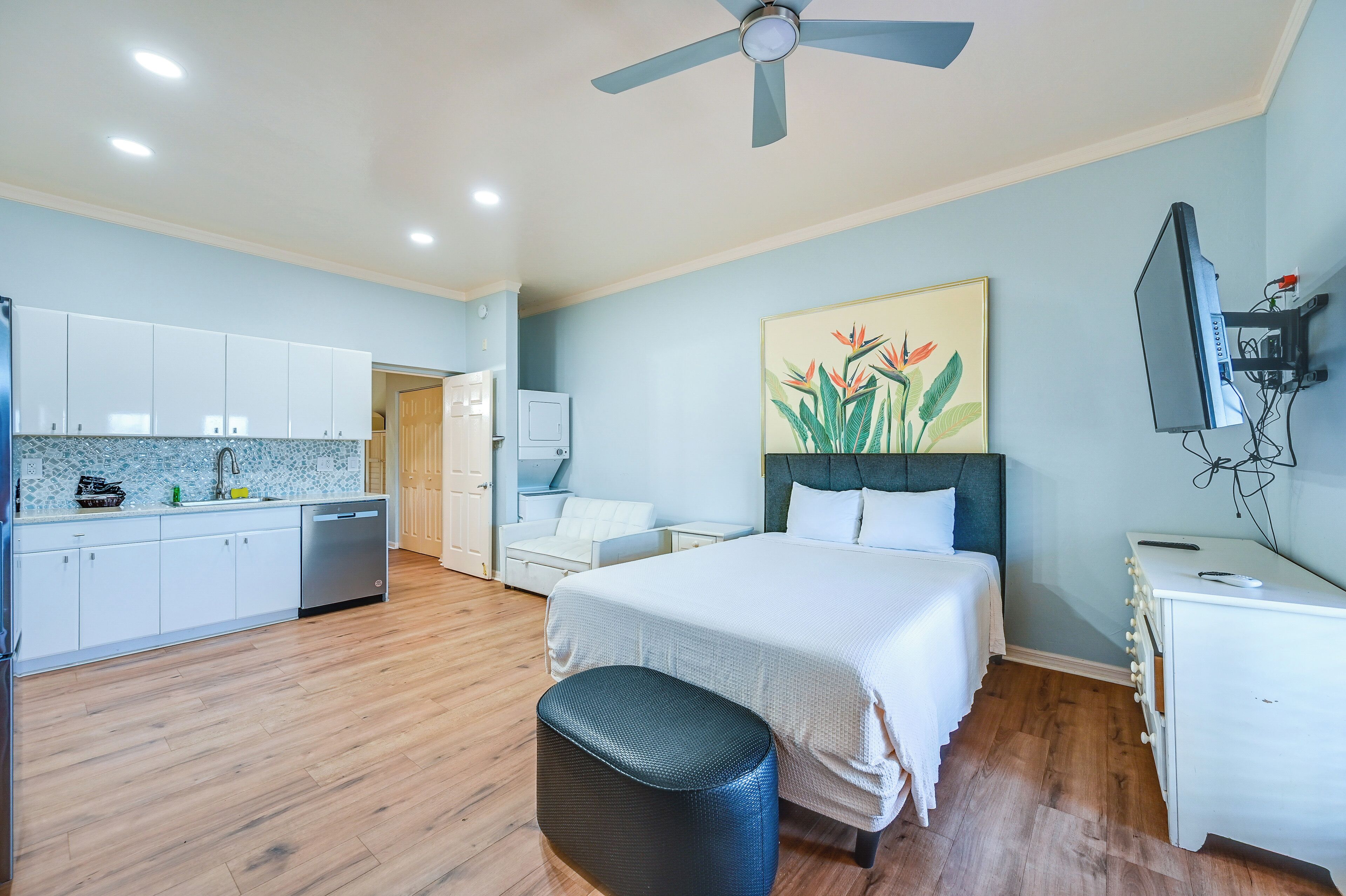Habitación de hotel con cama, escritorio, silla, televisor, ventilador de techo y una zona de cocina con armarios y fregadero.