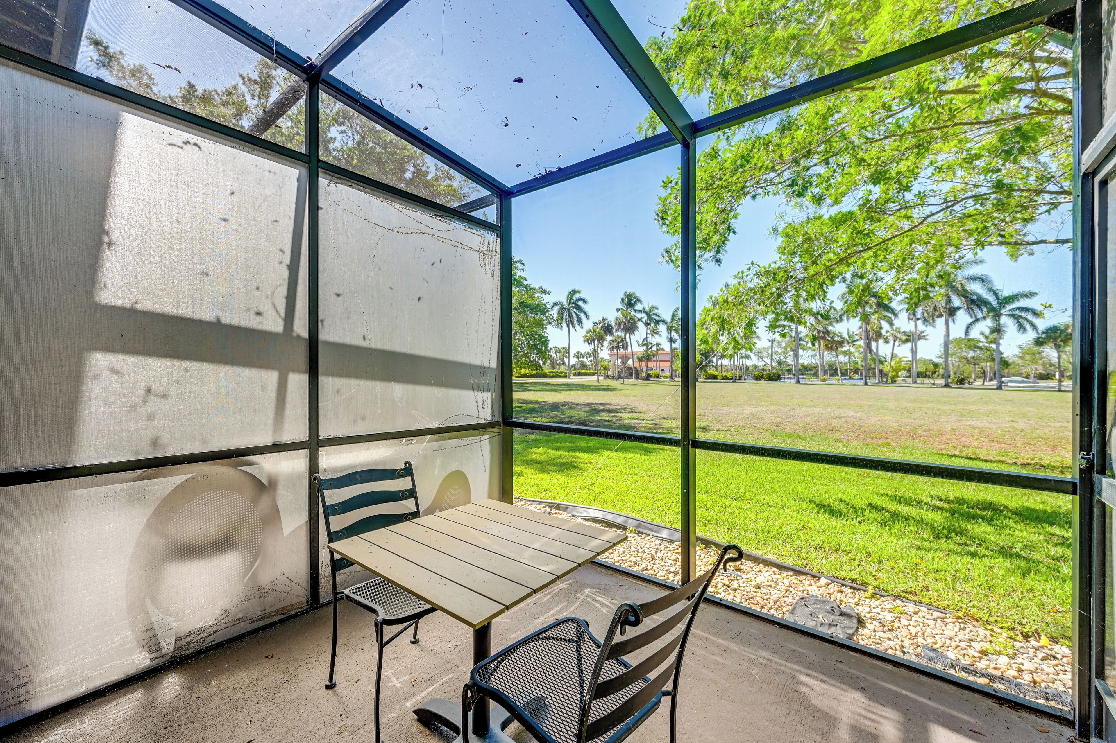 Un espace extérieur abrité, avec une table et des chaises, une grande fenêtre et une vue sur une zone verdoyante avec des arbres.