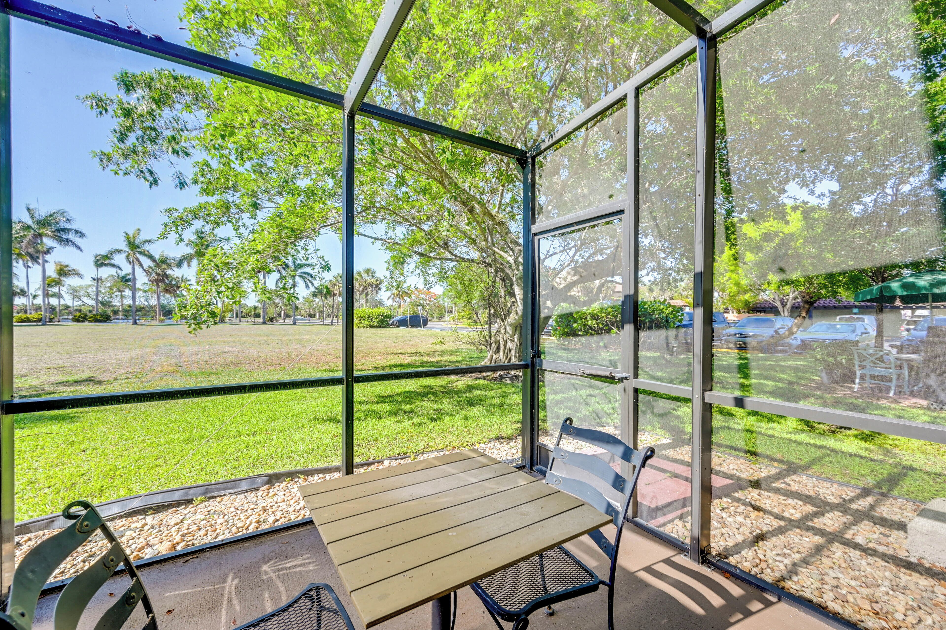 A covered outdoor seating area with a table and chairs, overlooking a grassy field and palm trees.