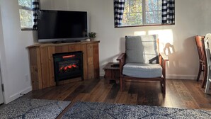 A living room with a fireplace, a television, a chair, and a window with checkered curtains. - Peace & Simplicity in a Little Red Cabin on the Farm (Lena)