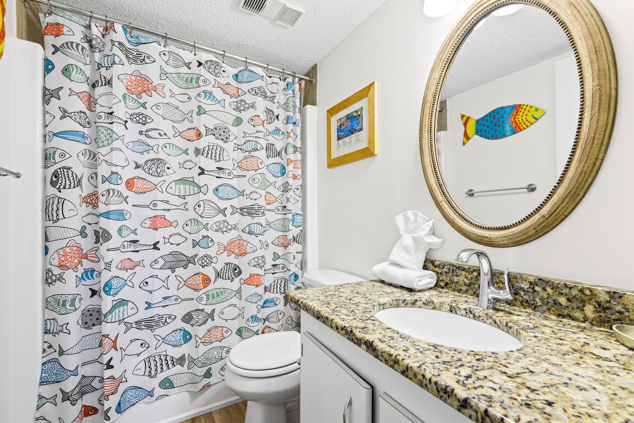Un bagno con tenda da doccia a motivi di pesce, un lavabo con piano in granito, un water bianco e uno specchio decorato con un pesce.