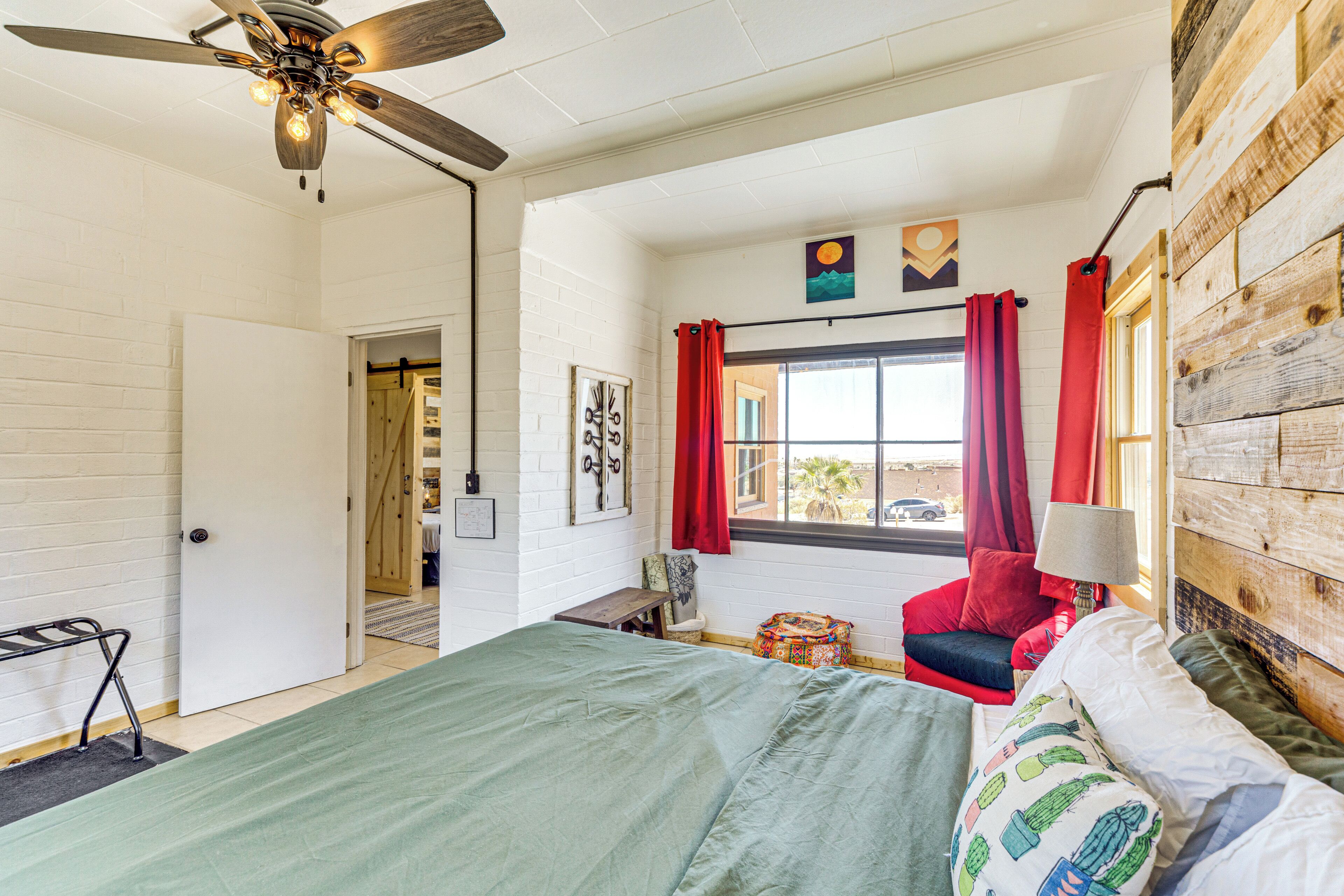 A bedroom with a bed, a ceiling fan, a window with curtains, a wooden wall, and a door.