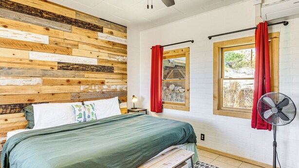 A bedroom with a wooden headboard, a bed with green bedding, a bench, a standing fan, and a window with red curtains. - 5 Mi to Joshua Tree National Park: Desert Oasis! (Twentynine Palms)