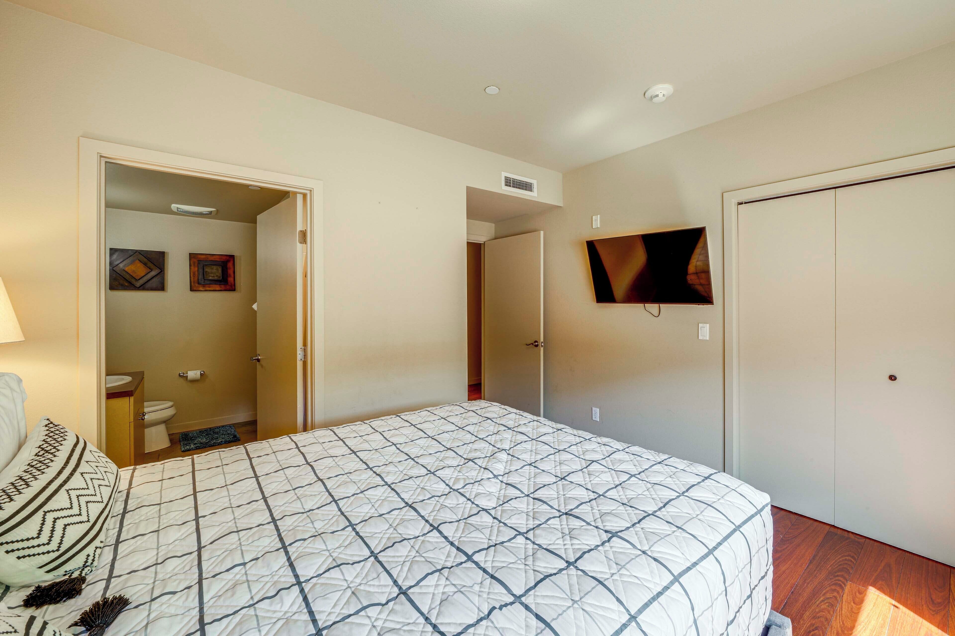 A bedroom with a bed, a TV mounted on the wall, a door leading to a bathroom, and a ceiling vent.