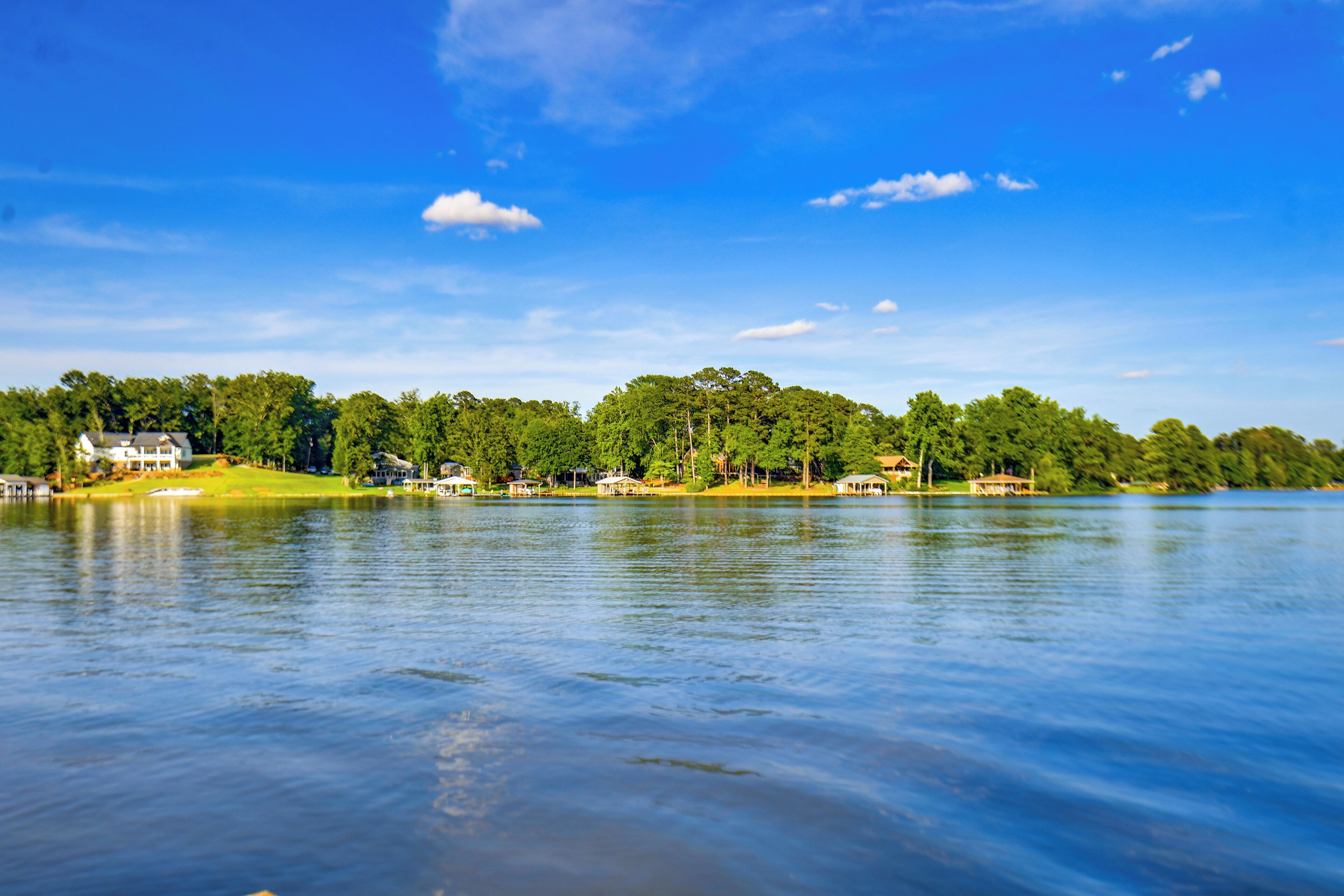 Un lago tranquilo con cielo despejado, rodeado de árboles verdes y algunas casas.