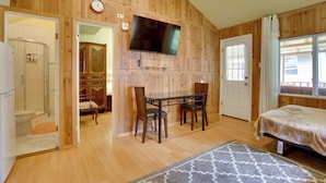 A wooden-paneled room with a bed, a TV mounted on the wall, a dining table with chairs, and a bathroom visible through an open door. - Screened Porch + Fire Pit: Cabin Near Bethel Woods (Mongaup Valley)