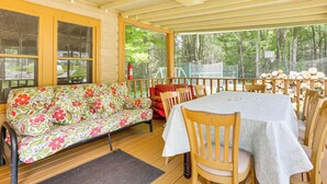 A covered porch with a wooden table and chairs, a floral sofa, and a view of the surrounding forest. - Community Perks: Monticello Cabin w/ Grill! (Mongaup Valley)