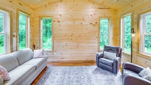 A cozy cabin interior with a wooden ceiling, a ceiling fan, a sofa, a chair, and large windows offering a view of the forest.