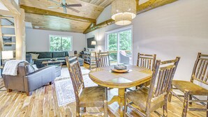A wooden dining table with chairs, a ceiling fan, and a chandelier in a room with wooden floors and large windows.